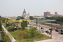slender yellow and light blue bridge across busy freeway with basilica and apartment towers in background