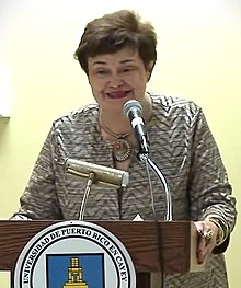 Woman with short hair in a geometrically printed blouse standing in front of a podium with a microphone and seal of the University of Puerto Rico on it.