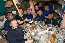 Nine[permanent dead link] astronauts seated around a table covered in open cans of food strapped down to the table. In the background a selection of equipment is visible, as well as the salmon-coloured walls of the Unity node.