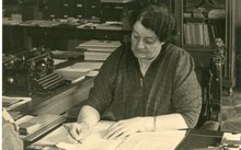 Photo noir et blanc d'une femme penchée en train d'écrire sur son bureau, un stylo à la main.