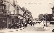 Photographie en noir et blanc représentant une place bordée de maisons et de cafés.