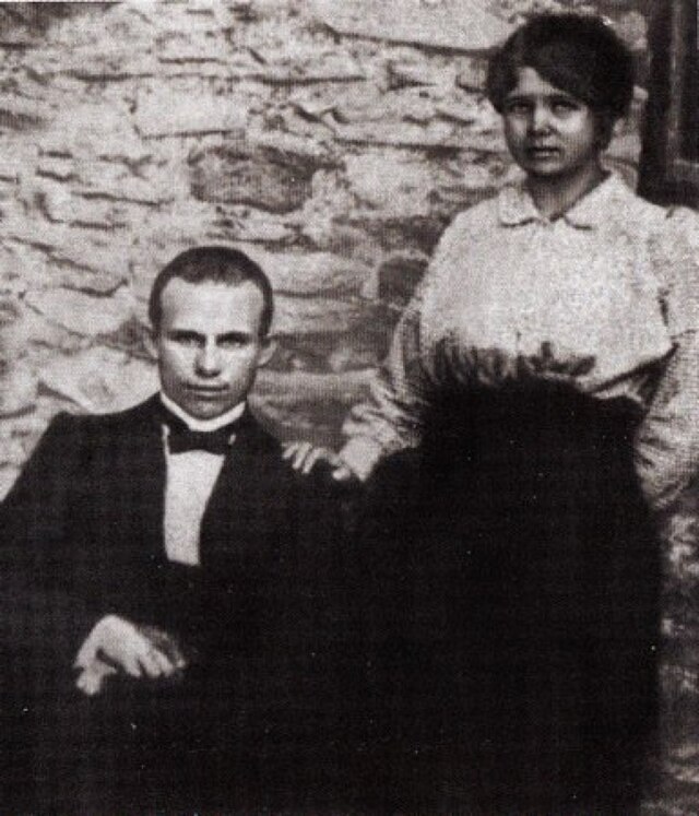 Photographie d'un homme en costume assis et d'une femme debout à côté de lui avec la main sur son épaule