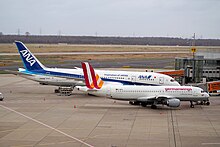 Photo de deux avions à la porte d'embarquement d'un aéroport. Au premier plan, un avion plus petit, aux couleurs blanches, oranges et rouges, cache la moitié d'un autre avion plus grand aux couleurs blanches et bleues, présent à ses côtés.