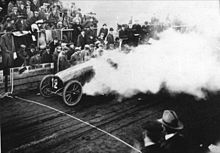 Photographie en noir et blanc d'une voiture automobile des années 1900 fumante devant la foule.