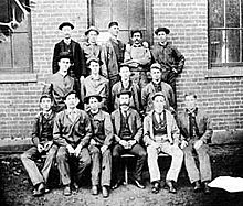 Групповое фото пятнадцати мужчин; пять в верхнем ряду, четыре в середине и шесть в переднем ряду, позируя перед кирпичным зданием.