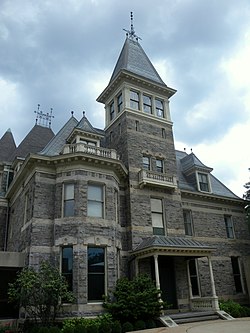 A stone house with a peaked-roofed tower in front and ornate roof and wood trim with a cloudy sky behind it