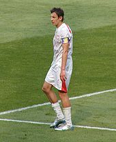 Portrait d'un joueur de football. Il porte un maillot blanc et est sur un terrain de football.