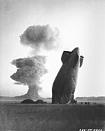 The tail, or “aft”, section of a U.S. Navy Goodyear ZSG-3 Blimp is shown with the Stokes cloud in background. The blimp was over five miles from ground zero when it was collapsed by the shock wave. The airship was unmanned and was used in military effects experiments on blast and heat. Navy personnel on the ground in the vicinity of the experimental area were unhurt. On the ground to the left are the re mai ns of the blimp's forward section.