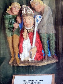 Painted statues of three men. The man in the centre is wearing a mitre and carrying a crozier and is staring straight forward. One of the two men flanking the central figure is carrying an axe.