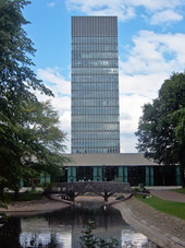 Blue/Grey skyscraper with a glass curtain wall. In the foreground is park pond with a small wooden bridge and trees at either side. In between the Tower and the pond is a modern 2-storey building