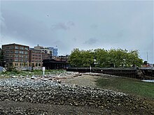 A stony beach overlooking several piers, brick buildings, and some trees