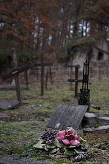 Vue du cimetière des Fous d’Évreux. En avant plan se trouve un objet funéraire représentant un bouquet de fleurs émaillées posé sur une sépulture. En arrière plan se trouve des croix de bois et le mur d'enceinte du cimetière ainsi qu'un petit bâtiment dégradé.