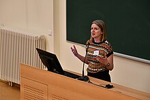 Dagmar Divjak teaching at the Summer School in Corpus Lingustics 2023, Tartu, Estonia. Divjak is standing behind a lectern with a computer on it and in front of a blackboard, and is gesturing animatedly.