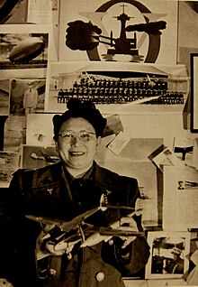 Sepia-Foto von Margaret Chung, die Modellflieger in beiden Händen hält. Sie trägt eine runde Brille und ihre schwarzen Haare als Victory Rolls, Lockenwickler-ähnliche Rollen. An ihrer Militärjacke ist am linken und rechten Schulterbereich je ein kleines, dreieckiges Abzeichen angebracht. Im Bauchbereich hängt zudem auf jeder Seite je ein rundes Abzeichen. Auf der Wand sind Fotos von Soldaten und verschiedenen Flugkörpern, unter anderem ein Kampfflugzeug und ein Zeppelin, zu sehen.