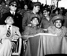 Trois hommes assis observent un événement. Le premier à gauche porte un costume et un fez tandis que les deux autres portent un uniforme et un casquette militaire. Trois hommes en uniforme se tiennent debout derrière eux.