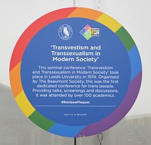 A circular commemorative plaque with a thick rainbow border. The logos of the Leeds Civic Trust and the Leeds LGBT+ Hub are visible near the top. The heading "'Transvestism and Transsexualism in Modern Society'" is written below. Below that is the following text in smaller print: "The seminal conference 'Transvestism and Transsexualism in Modern Society' took place in Leeds University in 1974. Organized by The Beaumont Society, this was the first dedicated conference for trans people. Providing talks, screenings and discussions, it was attended by over 100 academics." The two lines below, in smaller print, read "#RainbowPlaques" and "Supported by @StudioTDH".