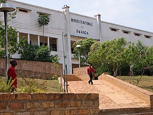 Blick auf einen über eine kurze Grünfläche führenden Weg mit einer Treppe am Ende vor dem Eingang des dreistöckigen Universitätsgebäudes. Über dem Eingang steht in Großbuchstaben Université nationale du Rwanda. Die unteren Stockwerke sind größtenteils durch Bäume verdeckt. Das erste Obergeschoss hat einen Laubengang und das Stockwerk darüber nur sehr kleine, schmale Fenster sowie ein Flachdach. Ein Mann kommt den Weg hinunter geschlendert.