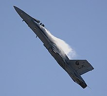 Jet fighter aircraft is seen against blue sky executing a pull-up, making it nearly vertical with contrail formed aft of the canopy.