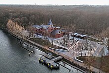 Ein längliches, dreistöckiges Backsteinhaus mit Giebeldach, umgeben von Wald, an der Spree. Im Vordergrund eine Schiffsanlegestelle auf Pontons im Wasser mit Sitzgelegenheiten und Geländern. Zwischen Anlegestelle und Haus sind zahlreiche Bänke und Tische im Freien als Sitzgelegenheiten.