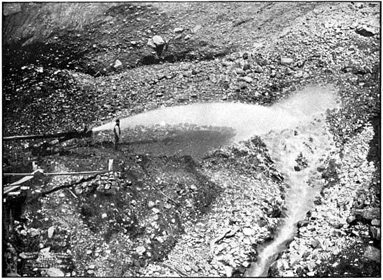 A person supervising a metal pipe shooting water against the soil; water is streaming down.