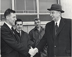 Diefenbaker, portant une veste par dessus son costume, serre la main d'un homme souriant. Deux autres hommes, semblant moins enthousiastes, sont à l'arrière-plan.
