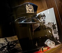 Une casquette, un insigne, un sifflet et deux photos de Louis de Funès dans une vitrine boisée.