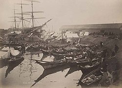 Canoes moored alongside a rocky beach dotted with tents, adjacent to a large ship and a warehouse