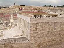 Крыша и верхняя часть колонного сооружения возвышаются над очень высокой каменной стеной с широкими пилястрами, украшающими верхнюю часть стены, и широким квадратом и ступенями у подножия стены.