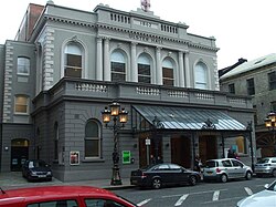 Photograph showing the front of Ulster Hall, a 19th-century grey and white rendered-brick building with black wrought-iron and glass canopy outside the front triple doors. Photo taken from the opposite side of a street, with cars parked on both sides.