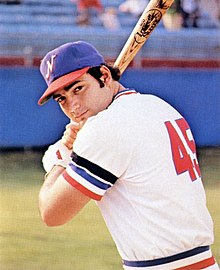 A man wearing a white baseball uniform with red and blue trim and a blue cap stands with his bat ready to swing.