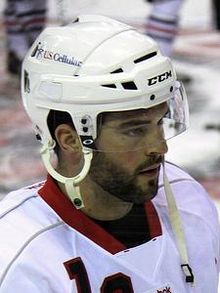 Photographie couleur d’un homme, barbu, de 3 quarts profil, portant un casque de hockey sur glace.