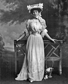 Old black-and-white photograph of a young white woman facing the camera, posed in front of a park bench and dressed in a long dress with a floral pattern, a small waist and a large hat