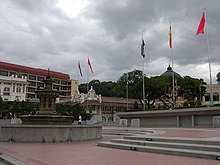 An ornate fountain at left with steps leading up to a wall with some of Malaysia's state flags on it.