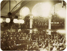 A photograph showing a large number of men seated on semi-circular tiers in a vaulted chamber as a large crowd looks on from an arcaded balcony