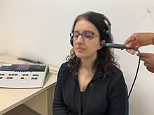 A imagem mostra uma mulher sentada em uma cadeira com uma sonda de imitanciometria em uma das orelhas e em uma mesa, ao lado da paciente, há o imitanciômetro no qual a sonda está conectada