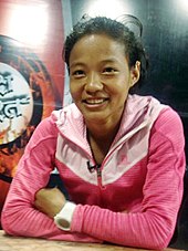 Mira Rai sitzt mit verschränkten Armen auf einen Tisch gestützt. Sie lächelt in die Kamera und trägt ihr schwarzes Haar zurückgebunden. Ihre sportlich aussehende Jacke ist rosa.