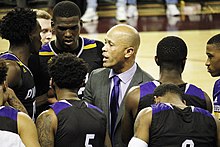 Photographie rapprochée d'un homme au crâne rasé parlant à des jeunes joueurs de basket-ball en maillot noir et violet.