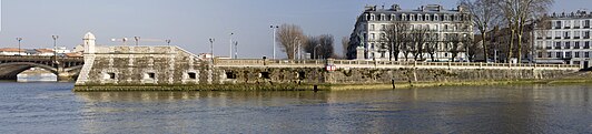 Vue d’un quai fortifié, orné d’une échauguette, en prolongement d’un pont sur un cours d’eau. À droite de la vue se dressent deux bâtiments imposants.