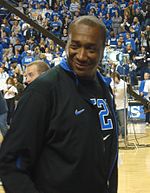 A man, wearing a black jacket and black T-shirt, is walking onto the basketball court.