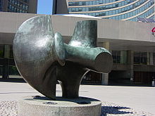Снимка на голяма бронзова абстрактна скулптура, пред стъклена и бетонна сграда.
