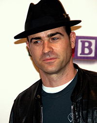 A black-haired man wearing a black fedora, a black leather jacket and a green T-shirt.
