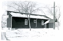 Black and white exterior photo of a single-storey rectangular wooden building with three windows on the longest side