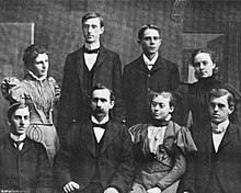 A black-and-white photograph of five men and three women, dressed formally, posing in front of a backdrop. The young Costello is tall and dark-haired. He is wearing a bowtie and eyeglasses.