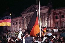 Farbfotografie in der Nacht von der Menschenmenge vor dem Reichstagsgebäude. Drei Deutschlandflaggen werden aus der Menge gehisst und nehmen einen Teil des Fotos ein. Auf den Stufen vor dem Gebäude steht auch eine Menschenmasse. Das Reichstagsgebäude ist beleuchtet.