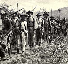 A column of Cuban Liberation Army soldiers marching. Most of the combatants are Afro-Cuban, wear torn clothing, and carry old rifles.