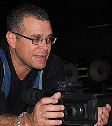 Against a dark background, Daranas leans forward on a surface while speaking and holding a digital camera.