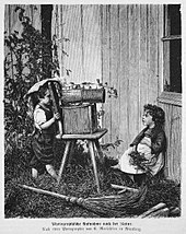 Eine Schwarzweißzeichnung von einem kleinen Jungen und einem kleinen Mädchen, die im Garten sind. Das Mädchen sitzt angelehnt an einem Holzhaus und der Junge schaut hinter einem Stuhl durch eine Röhre, die auf einem Tierkäfig aus Holz liegt.