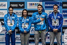 Ferraioli trägt eine blaue Trainingshose, die anderen drei eine graue. Der Haltung der Vier ist zu entnehmen, dass dieses Foto beim Abspielen der Nationalhymne entstanden ist.