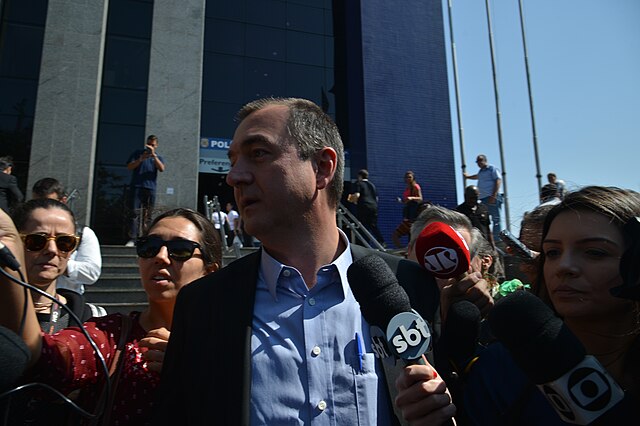 Um homem branco, trajando uma camisa azul e terno preto, é cercado por jornalistas. Uma entrevistadora do SBT põe o microfone na frente de Joesley.