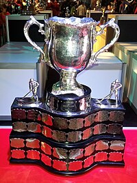 A silver cup attached to a large black base with several medallions each engraved with the name of the team and players for each successive champion.